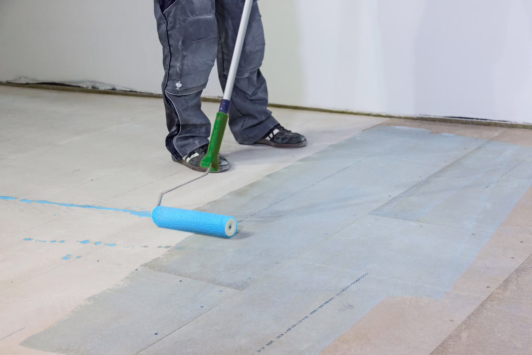 UZIN_Application Image_PE360 Plus_Renoplan System_2020-10 On voit les jambes et les pieds inférieurs d'un ouvrier qui applique un primaire ou une peinture bleu clair sur un sous-plancher en bois à l'aide d'un rouleau à long manche. La personne porte un pantalon de travail foncé et des baskets noires ; le rouleau laisse un film bleu humide sur les lames de plancher dans une pièce intérieure aux murs blancs. L'image met l'accent sur la préparation du sol, les outils et les éclaboussures de peinture.