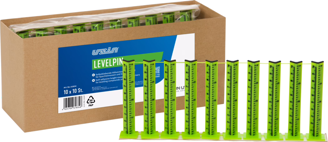 UZIN_Dummy_Levelpin_2026-02 Une boîte en carton contenant plusieurs languettes vertes vives autocollantes pour nivellement ; dix onglets de mesure verticaux fixés sur une base et reliés par un fin cordon jaune sont présentés au premier plan, avec une étiquette produit bleu et blanc portant l'inscription 'LEVELPIN'.