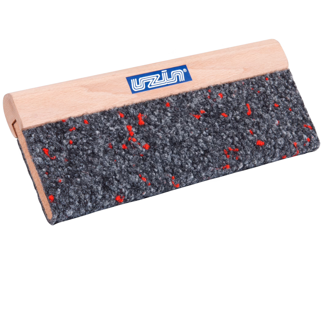 Gros plan d'un outil rectangulaire avec une poignée en bois clair et un patin en feutre gris grossier parsemé de petites fibres rouges ; une petite étiquette de marque bleue est visible sur la poignée en hêtre lisse. L'outil est photographié en angle sur fond blanc, mettant en valeur la texture du feutre et le grain fin du bois.