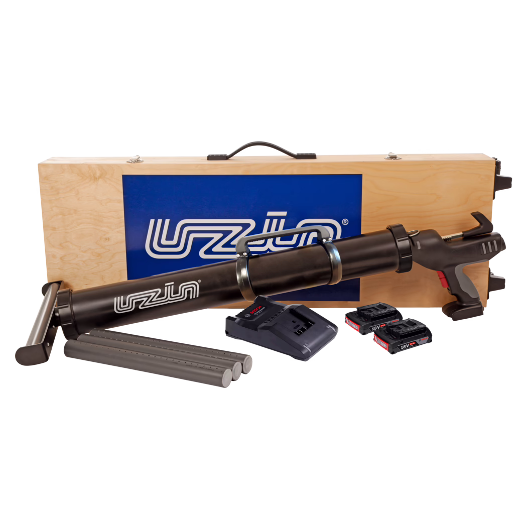 UZIN_Dummy_ATD 100 NEW_2021-04 Mallette de transport en bois avec un pistolet à cartouche sans fil noir et ses accessoires : on distingue le dessus d'une mallette en contreplaqué clair avec une poignée noire et un panneau logo bleu, derrière un long pistolet à cartouche noir alimenté par batterie. Devant la mallette se trouvent trois cartouches grises, une base de chargeur et trois accus lithium-ion 18V. Le pistolet présente une poignée pistolet avec des accents gris et rouges, des colliers métalliques autour du canon de cartouche et une gâchette. Composition de studio sur fond blanc.